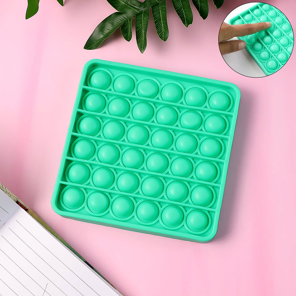 Overhead view of a square green silicone pop-it fidget toy on a pink surface with green leaves in the background.