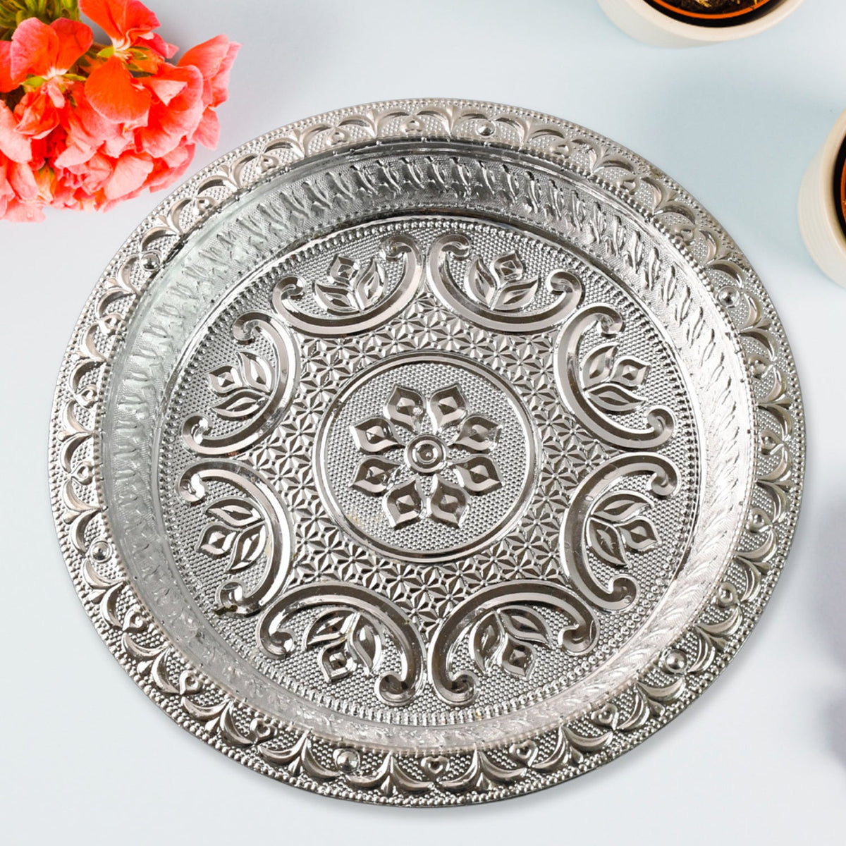 A silver-colored plastic pooja thali with embossed patterns, surrounded by orange flowers and two white containers on a