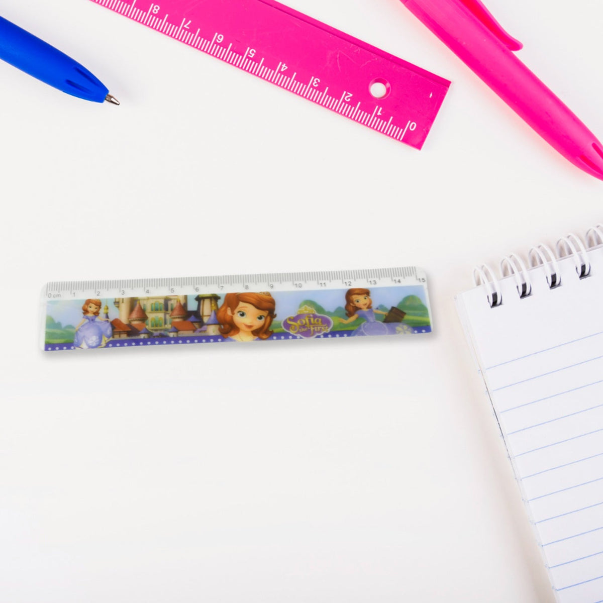 A ruler on a white desk next to a spiral notebook, with a blue pen and pink ruler in the background.