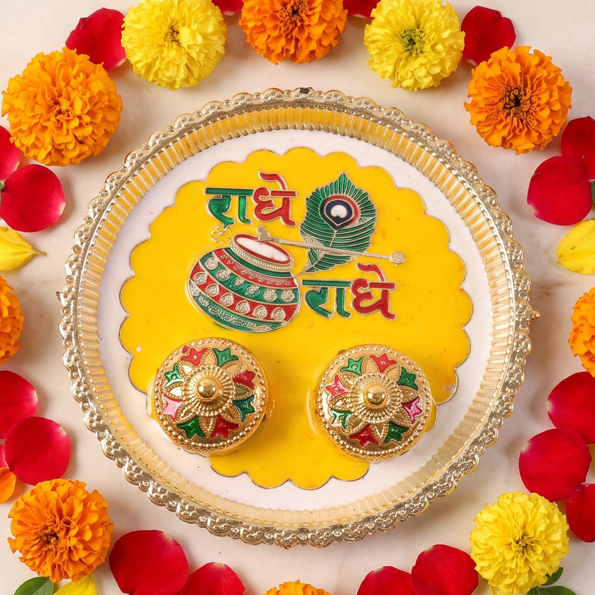 A silver and yellow Radhe Radhe pooja thali set with two small decorative containers, surrounded by marigolds and rose