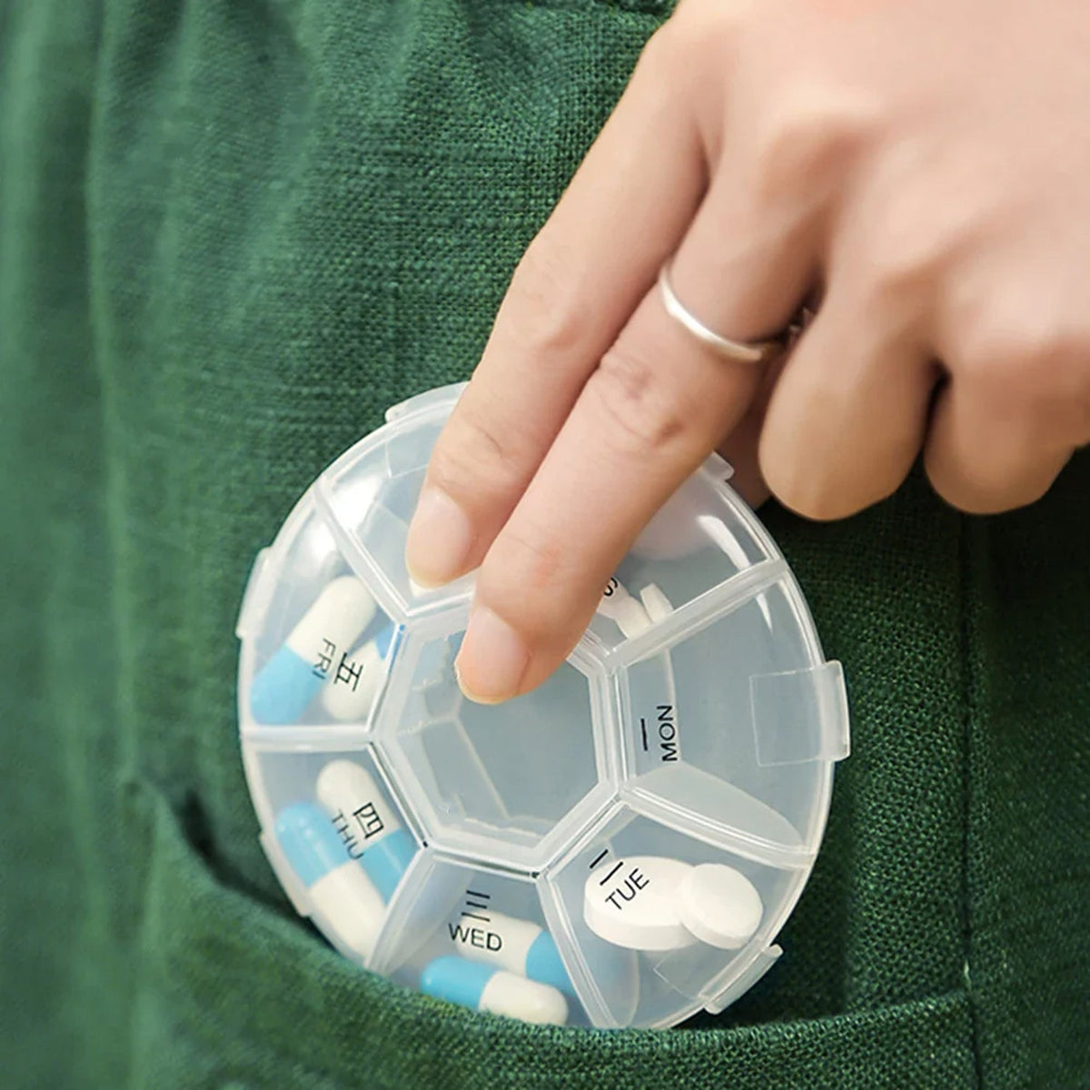 A hand placing a clear 7-day pill organizer with blue and white pills into a green pocket.
