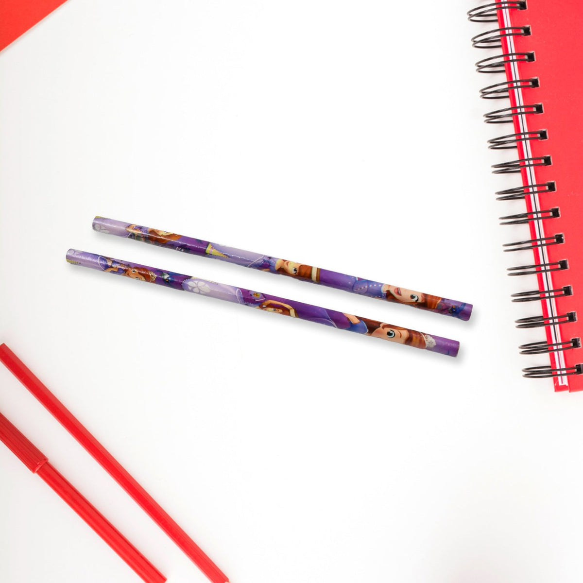 Two purple pencils lying on a white surface next to a red spiral notebook and red pens.