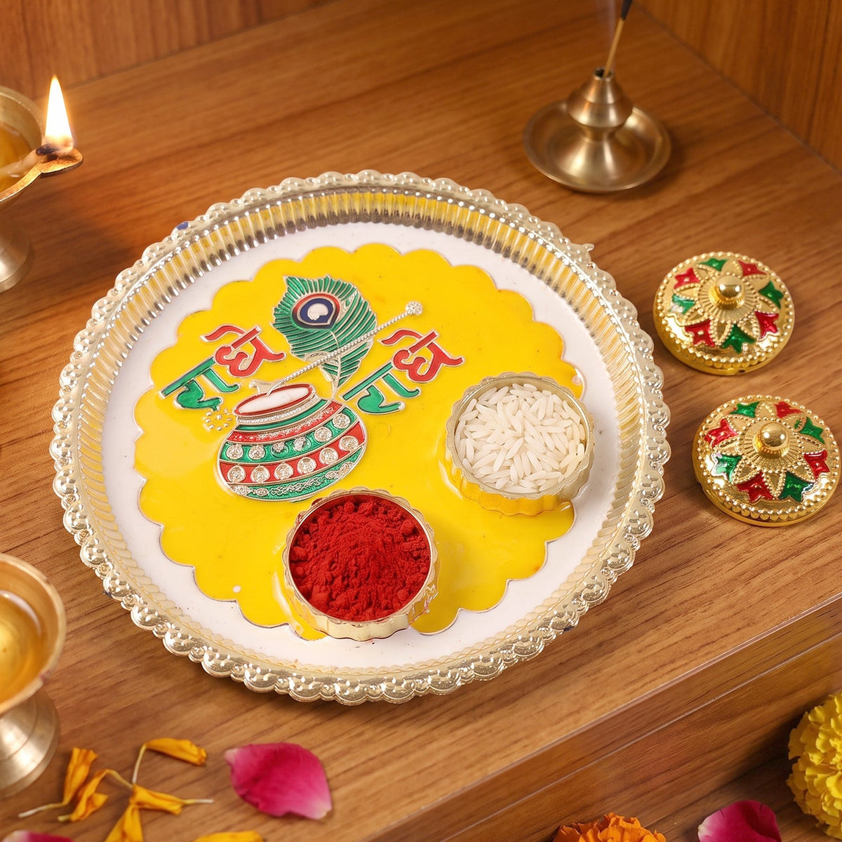 A silver and yellow Radhe Radhe pooja thali with small containers of kumkum and rice, next to two decorative containers.
