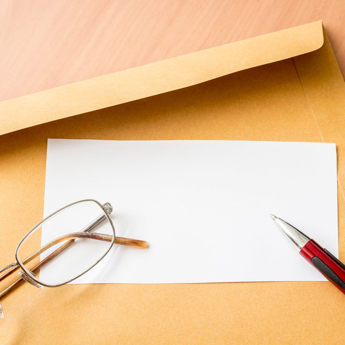 A close-up of a brown kraft envelope with a blank white card, reading glasses, and a pen on top.