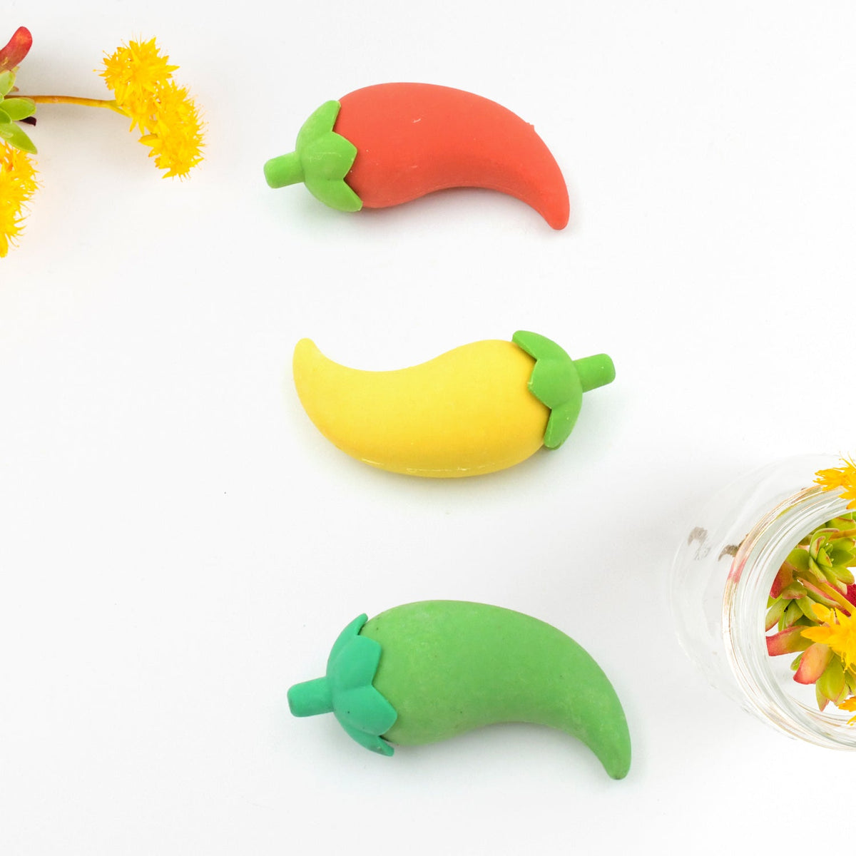 Three chili pepper erasers in red, yellow, and green, arranged vertically on a white surface with yellow flowers.