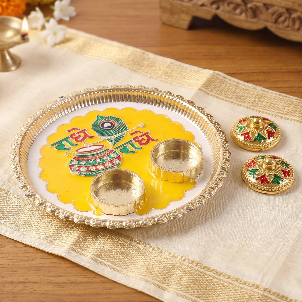 A silver and yellow Radhe Radhe pooja thali with empty containers, next to two decorative containers on a white cloth.