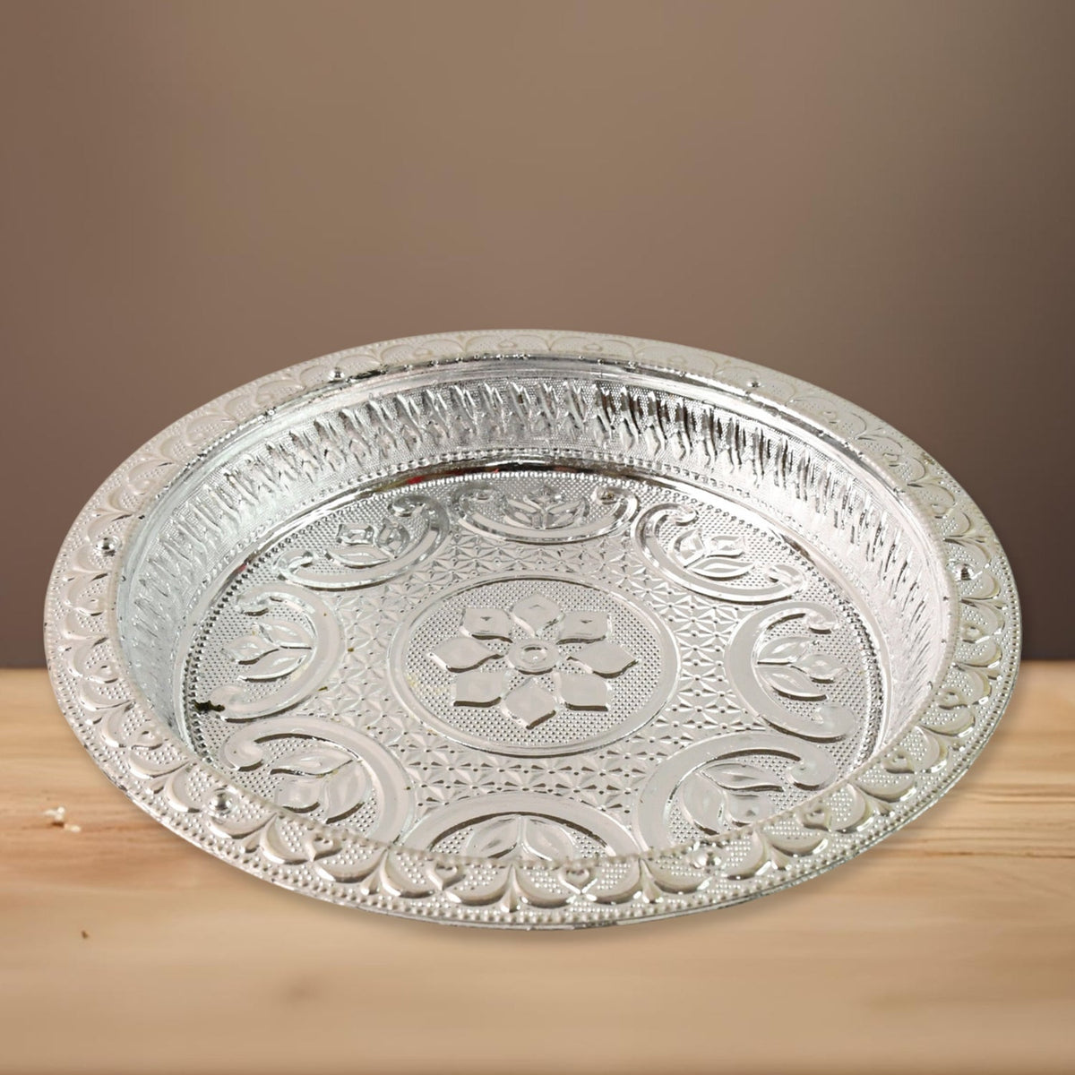 A silver-colored plastic pooja thali with embossed floral designs, resting on a light wooden surface with a brown background.