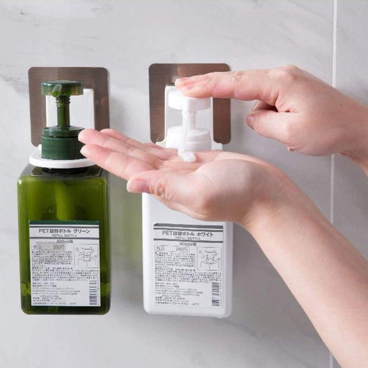 A person dispenses white lotion from a white bottle in a wall-mounted holder, next to a green bottle in another holder.