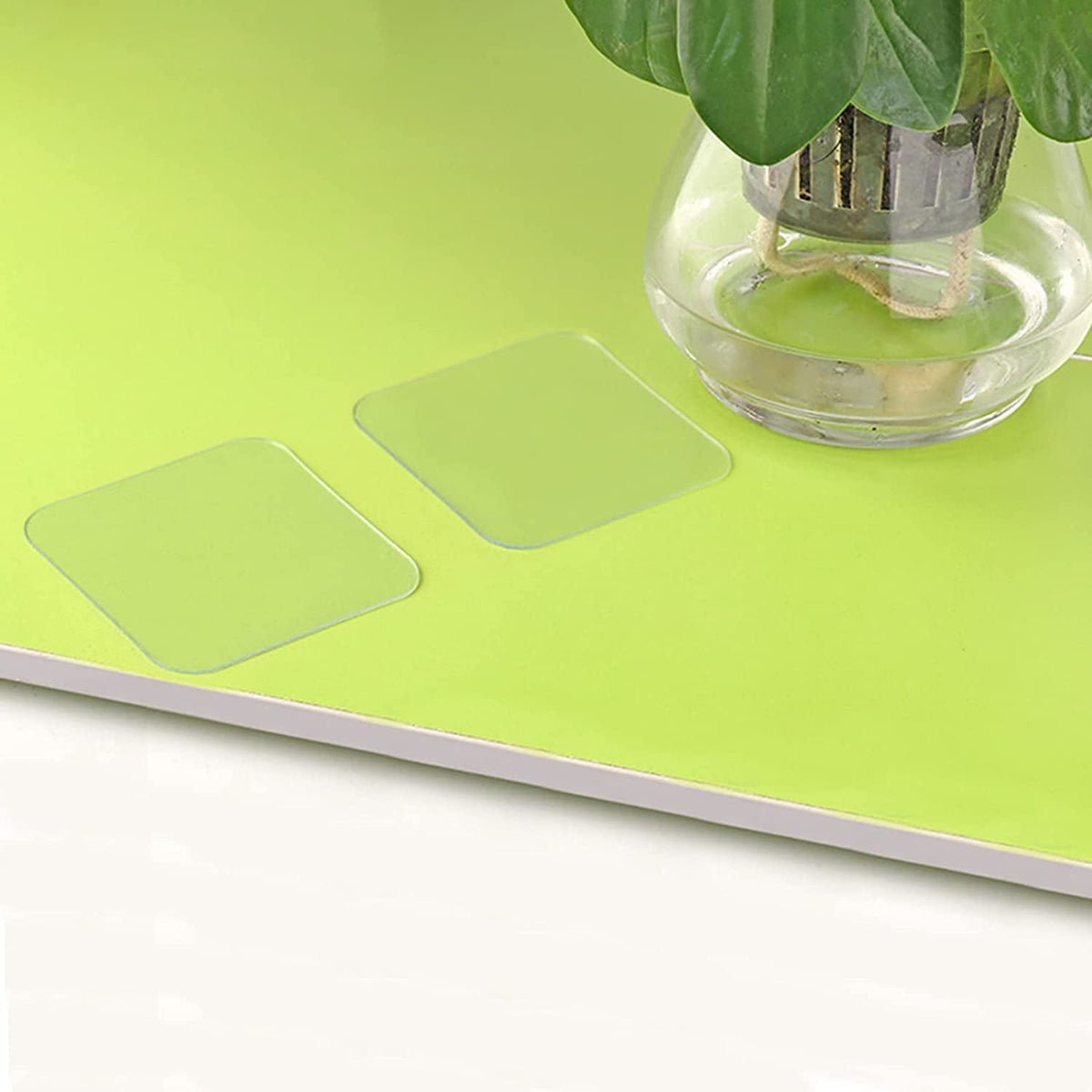 Two clear adhesive squares are shown on a bright green surface next to a glass vase with a plant.