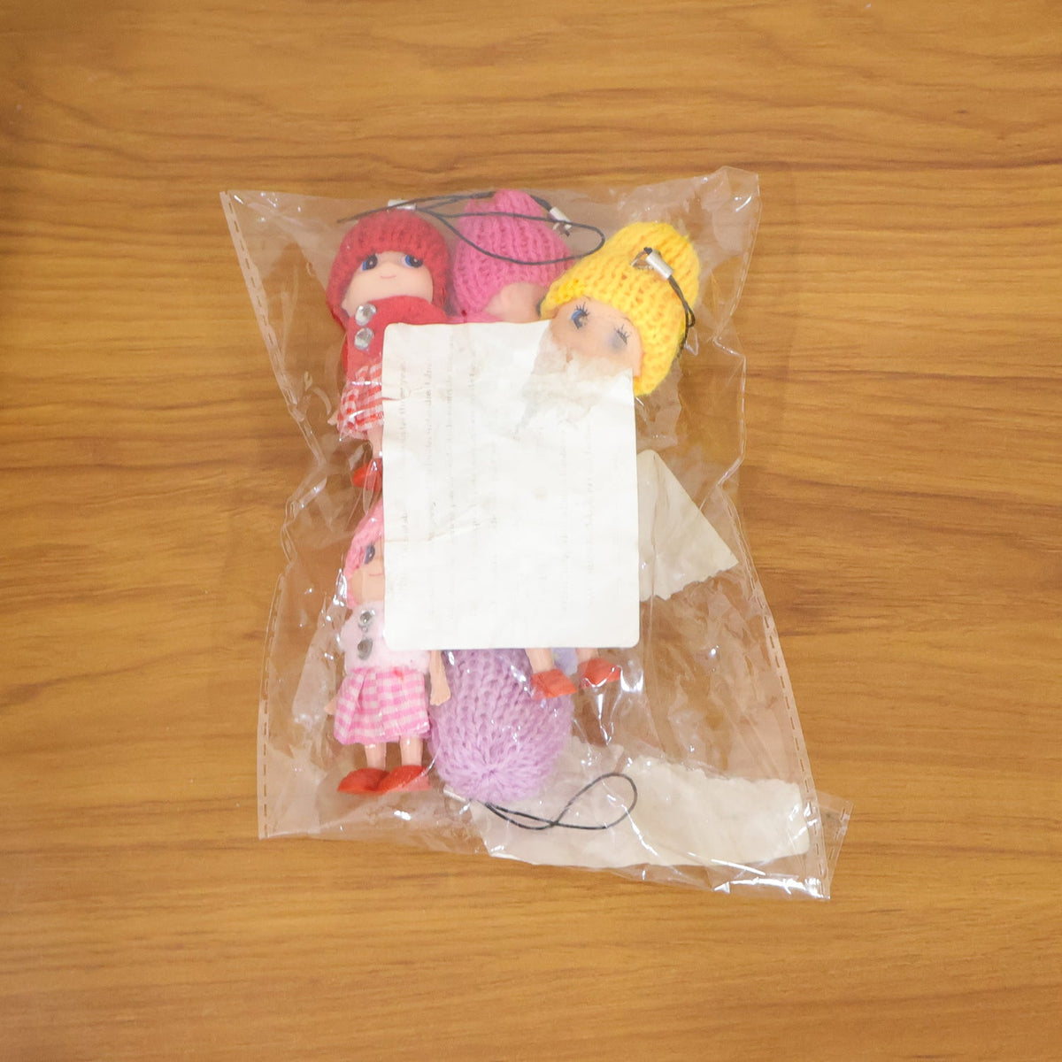 A clear plastic bag containing several colorful doll keychains with knitted hats is on a wooden surface.