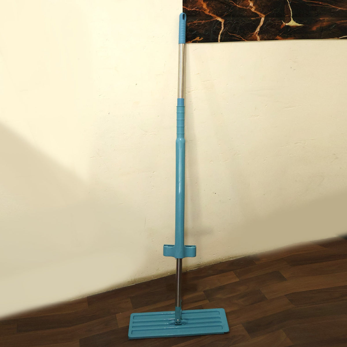 A blue and silver flat mop with a grey microfiber pad stands upright against a light wall and dark wooden floor.
