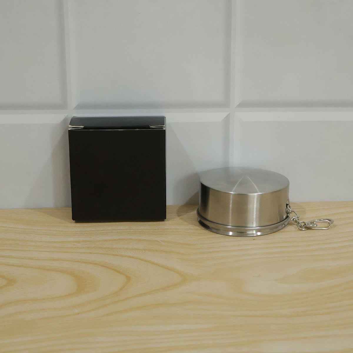 A black box and the closed stainless steel collapsible cup with a keychain are on a light wooden surface against a white