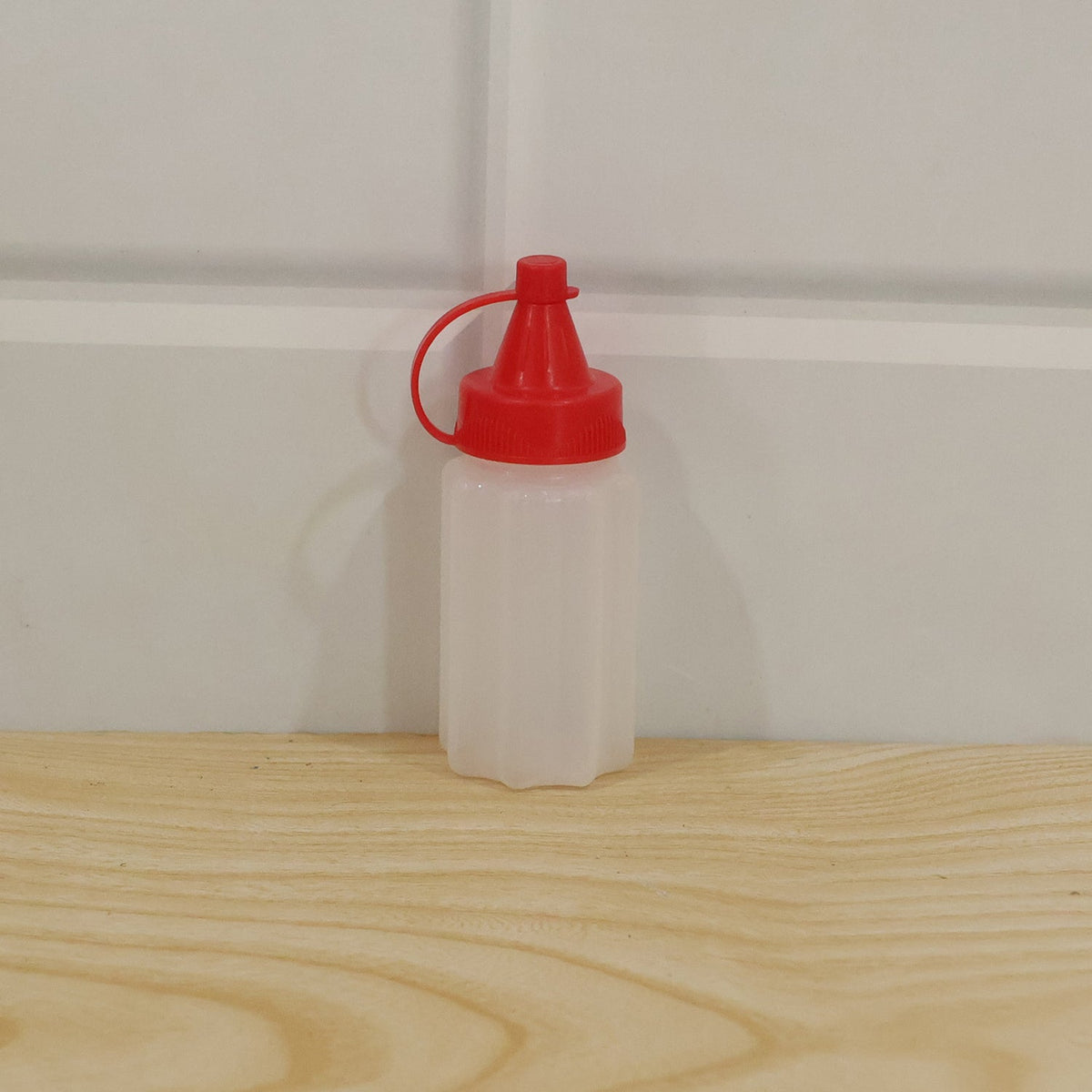 A small, empty, white ribbed plastic squeeze bottle with a red cap sits on a light wooden surface against a white wall.