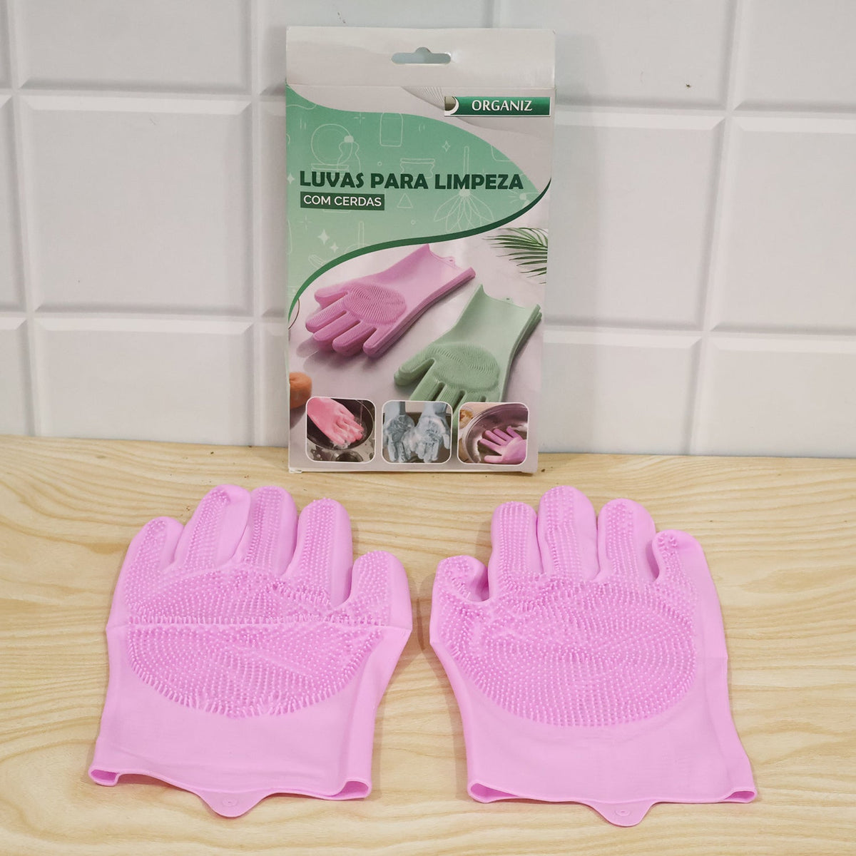 A pair of pink silicone scrubber gloves resting on a wooden surface in front of a product box for "Luvas Para Limpeza Com