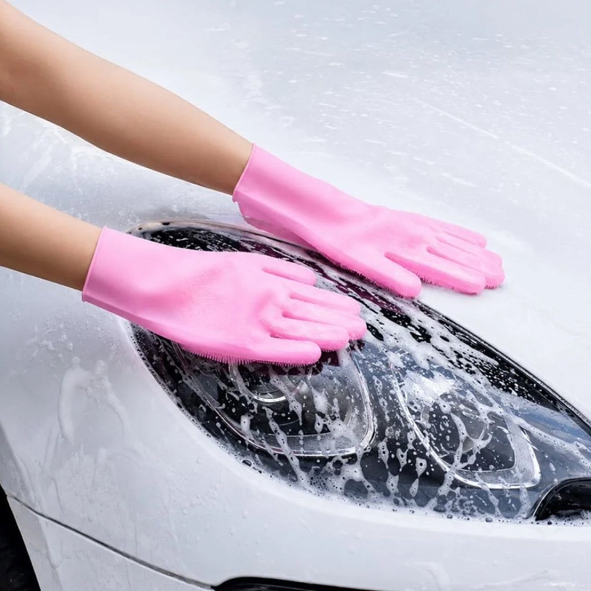 A person wearing pink silicone gloves with integrated scrubbers, cleaning the hood of a white car covered in suds.