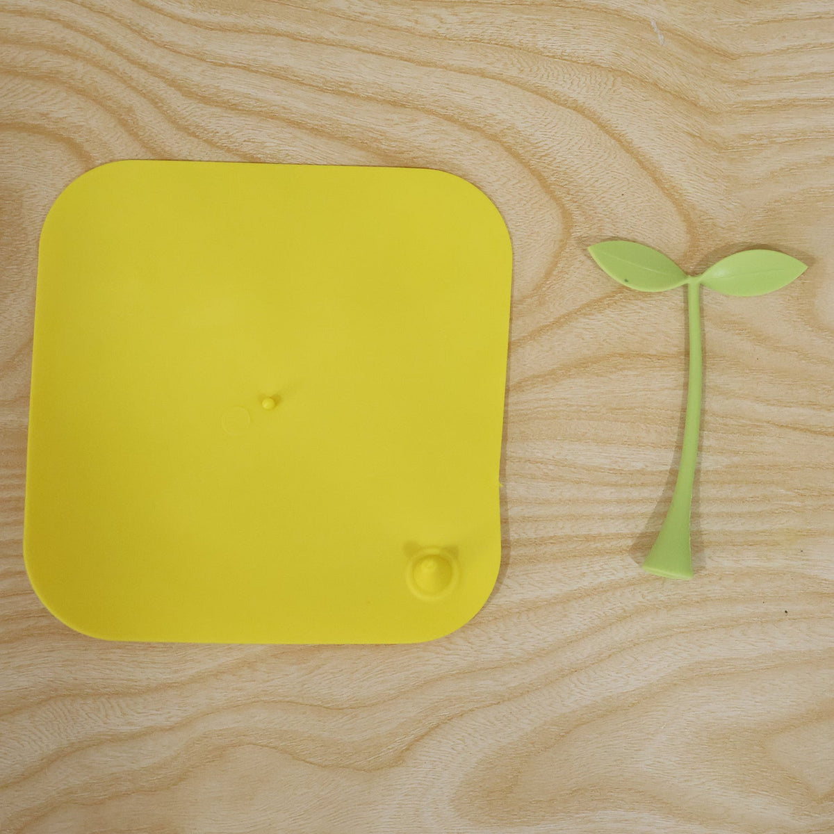 A bright yellow square silicone drain cover and its detached green sprout-like handle on a light wooden surface.