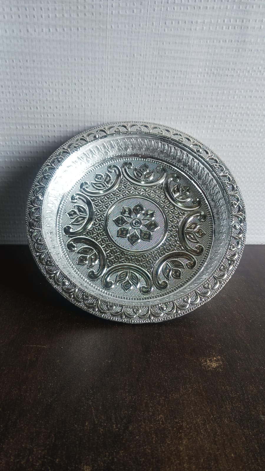 A silver-colored plastic pooja thali with embossed patterns, on a dark brown surface with a white textured wall in the