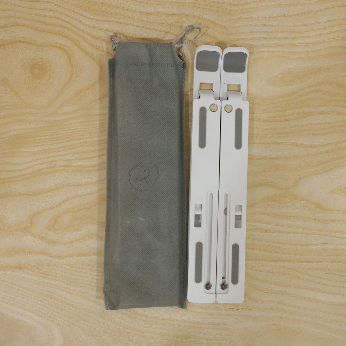 White folded laptop stand next to a gray fabric carrying pouch, both resting on a light wooden surface.