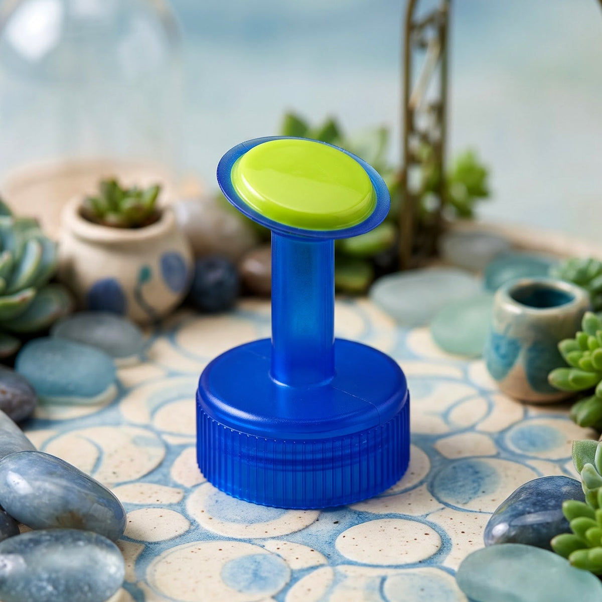 A blue and green plastic sprinkler cap stands on a patterned blue and white surface surrounded by small succulents and