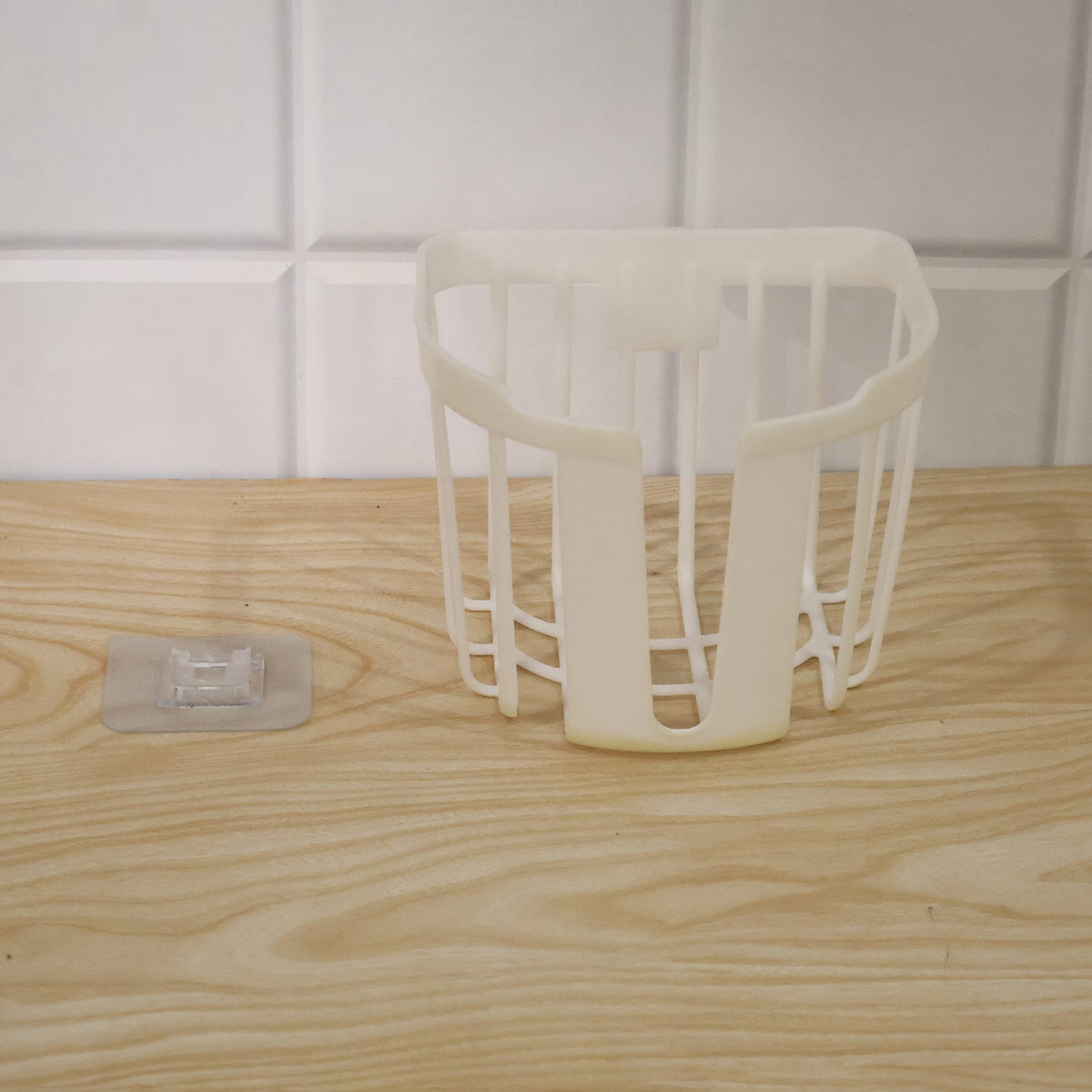 A white plastic toilet paper holder sits on a wooden surface next to a clear adhesive hook, with white tiles in the