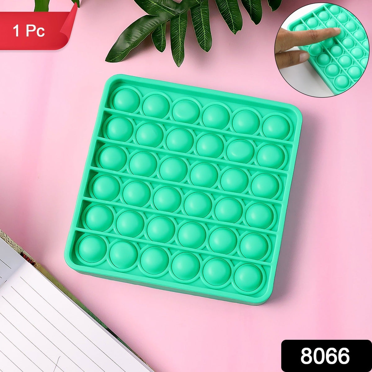 Overhead view of a square green silicone pop-it fidget toy on a pink surface with a plant and notebook.