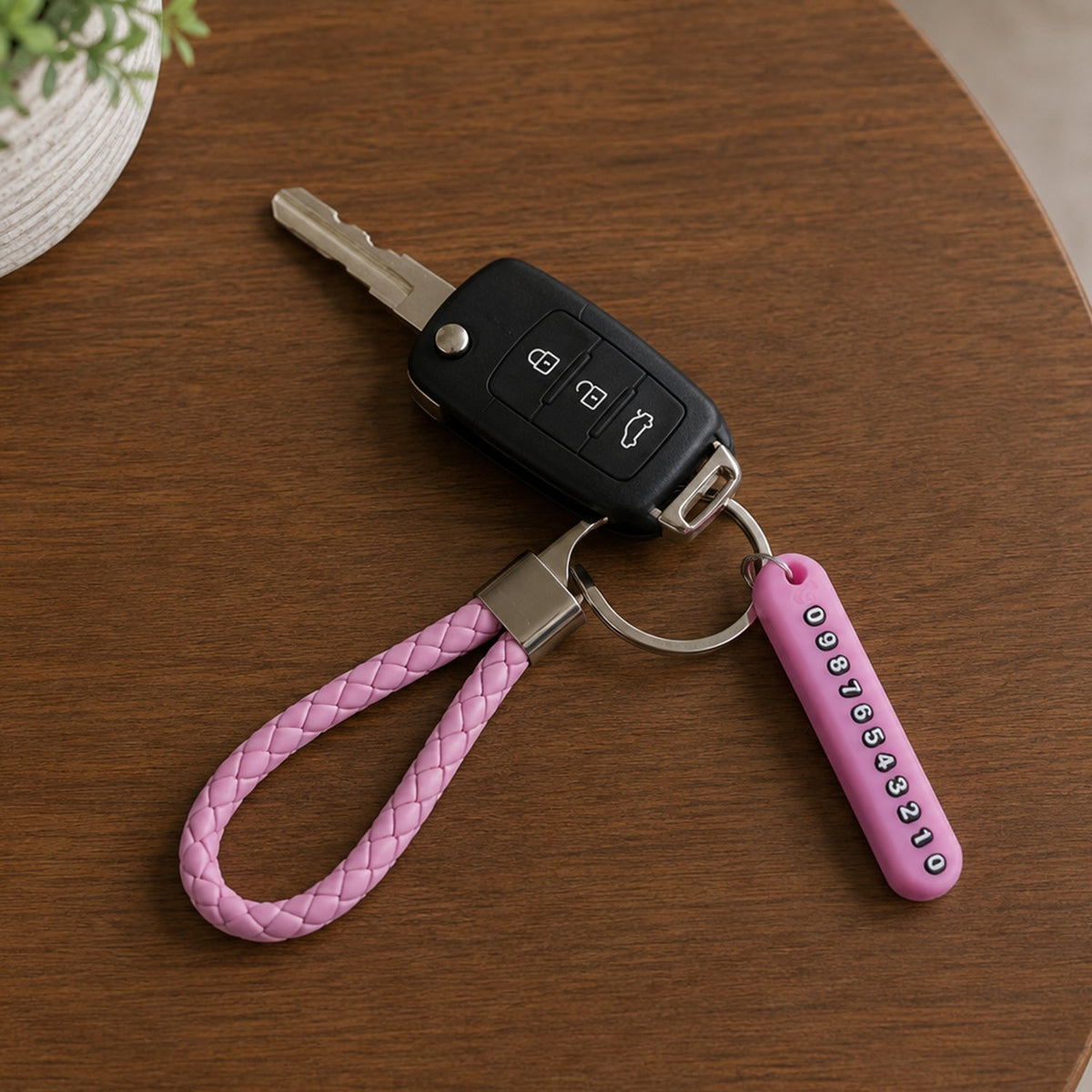 A pink braided wrist loop keychain with a car key and a pink number tag on a wooden table, with a plant in the background.