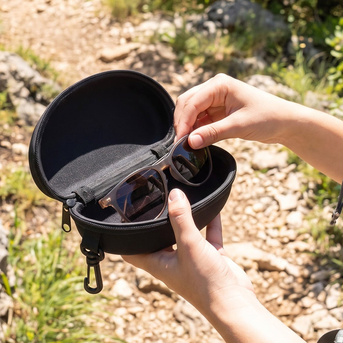 Hands placing brown sunglasses into an open black hard shell case with a clip, outdoors on a rocky path.