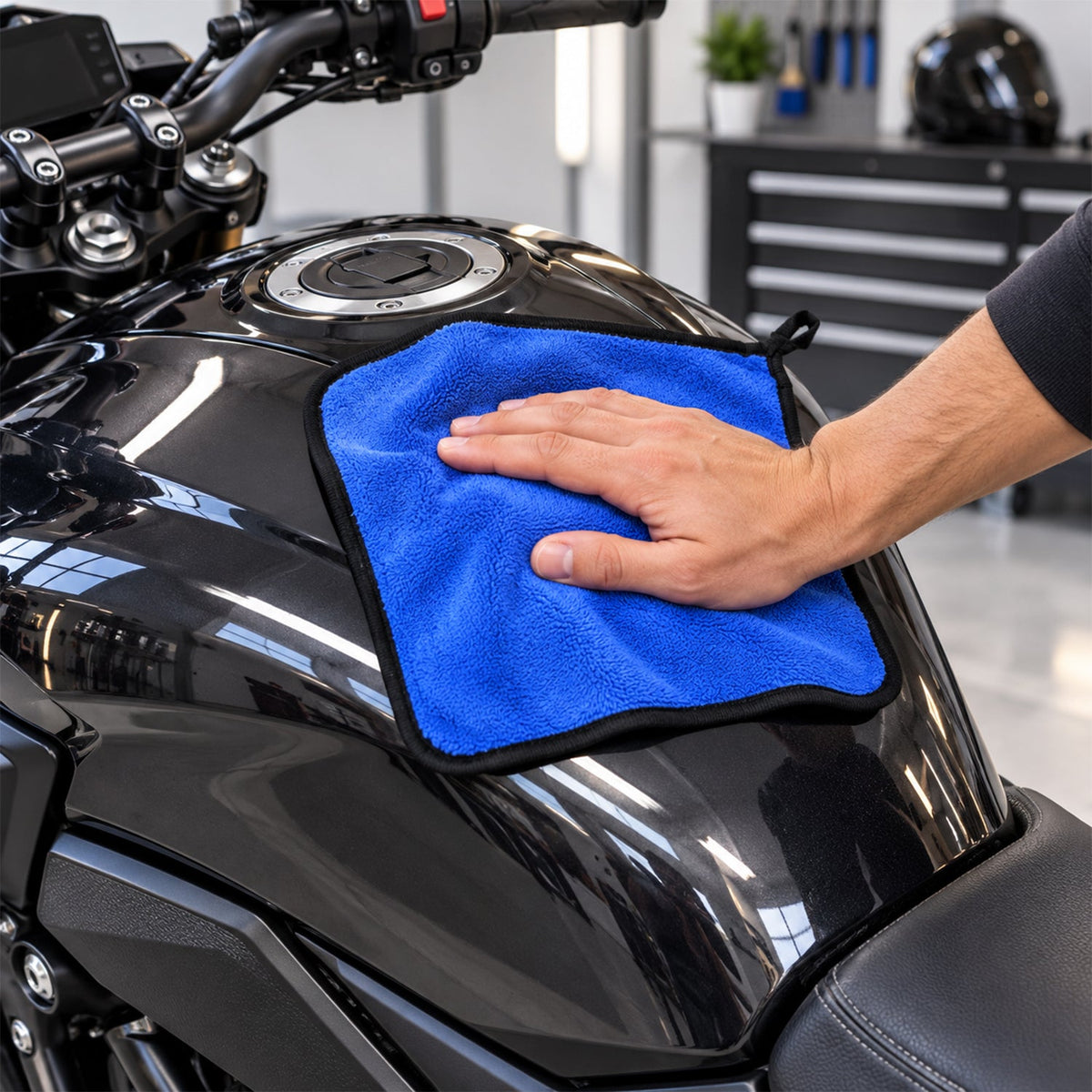 A hand uses a blue microfiber cloth to clean the shiny black fuel tank of a motorcycle.