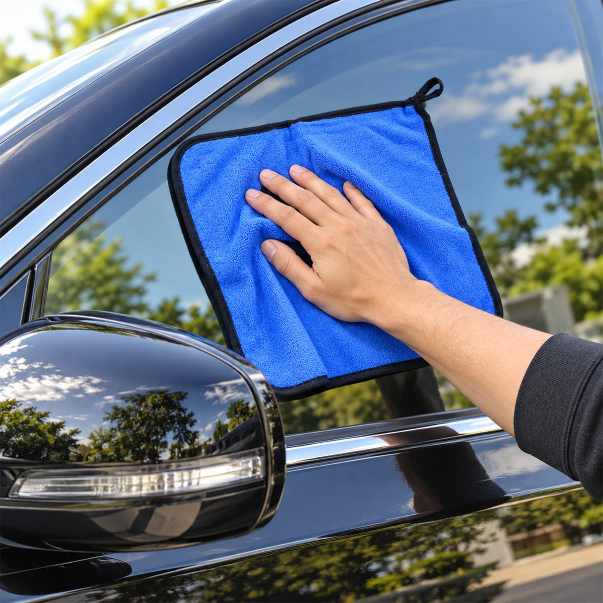 A hand uses a blue microfiber cleaning cloth to wipe the window of a black car, with a side mirror visible.