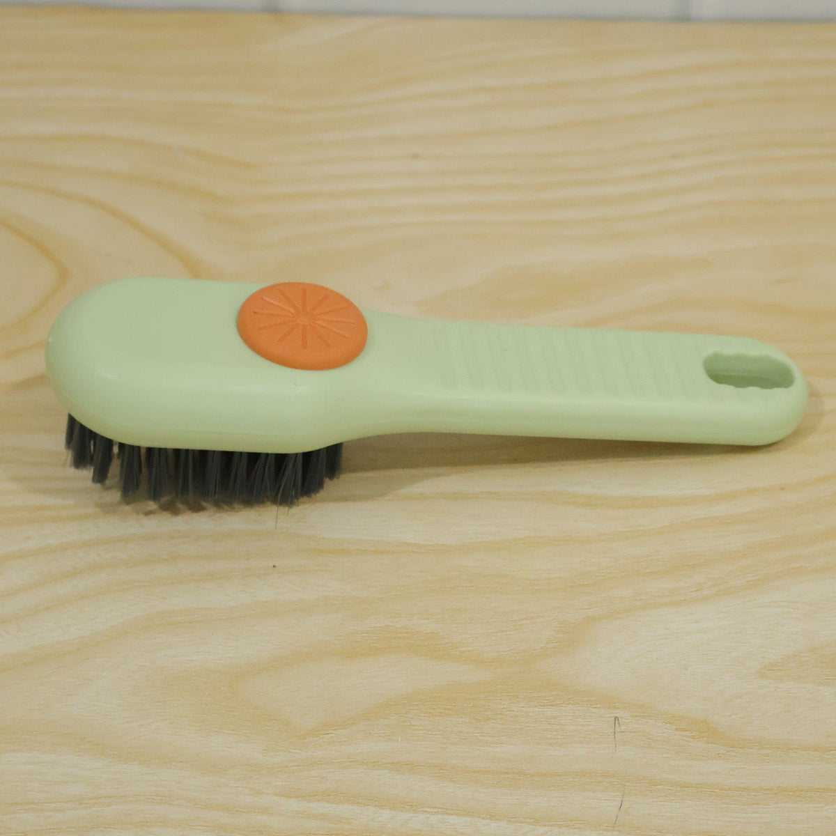 A green and orange liquid-dispensing brush with dark bristles is positioned on a light brown wooden surface.