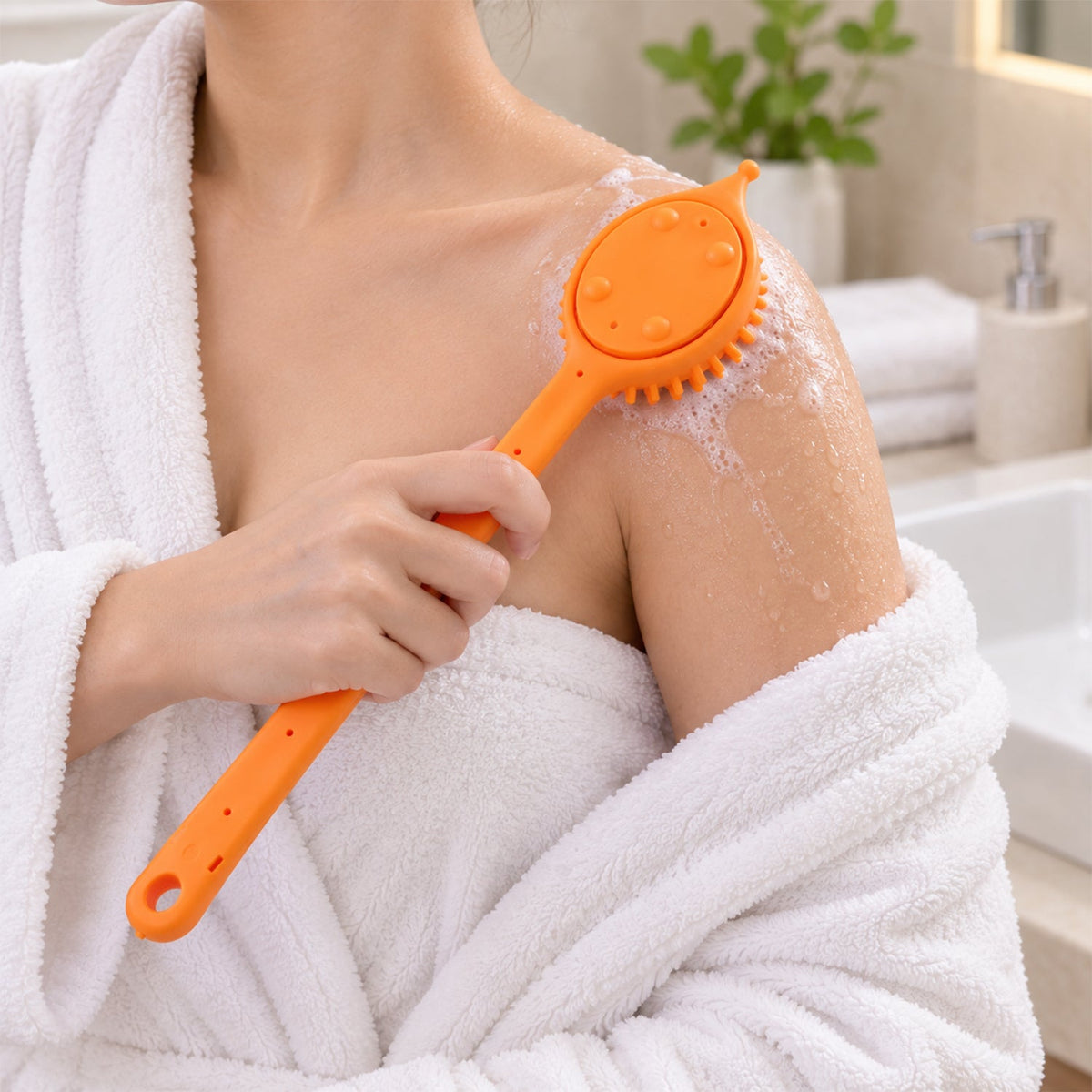 A person in a white robe uses an orange silicone back scrubber with soap suds on their shoulder in a bathroom.