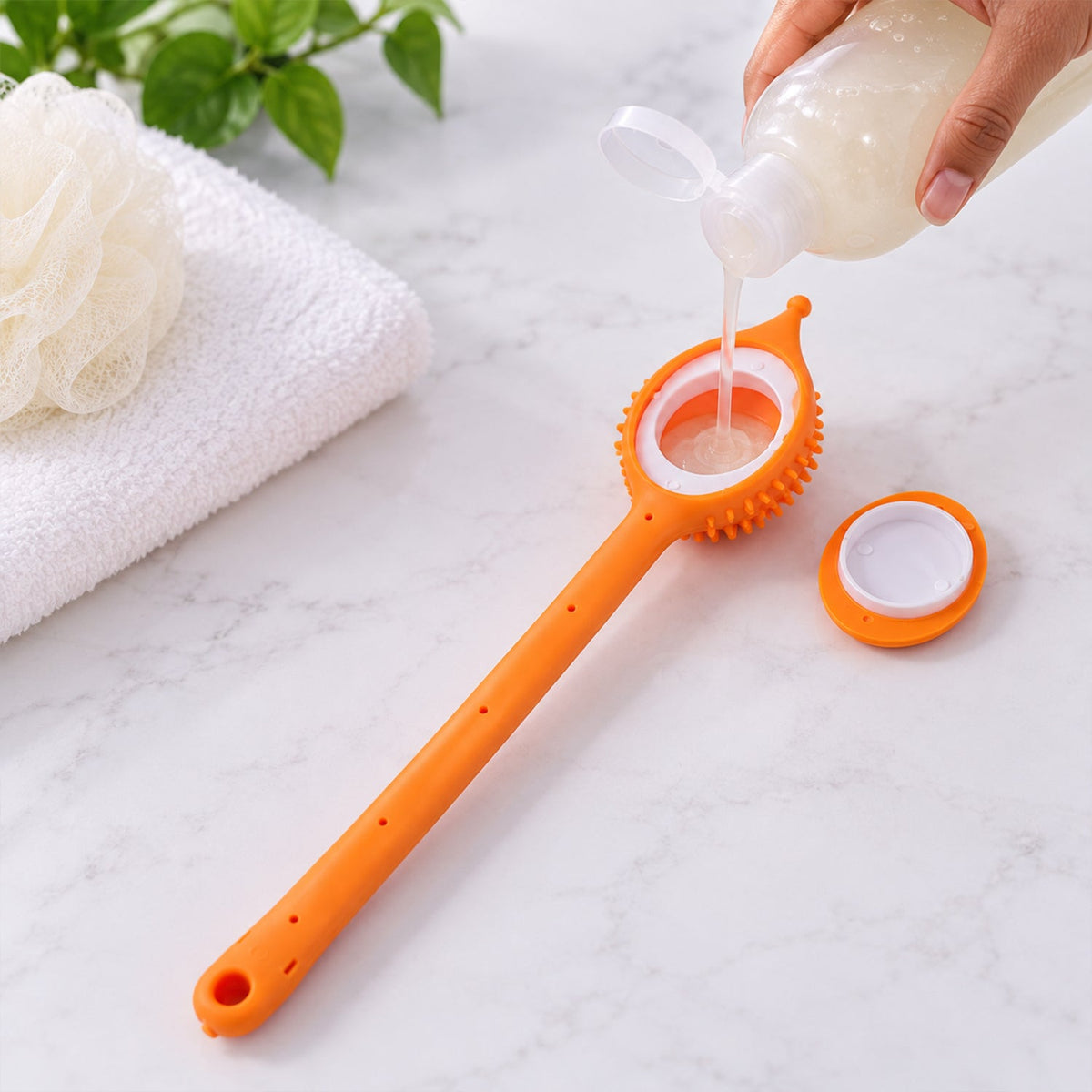 A hand pours liquid soap into the open compartment of an orange silicone back scrubber on a marble counter.