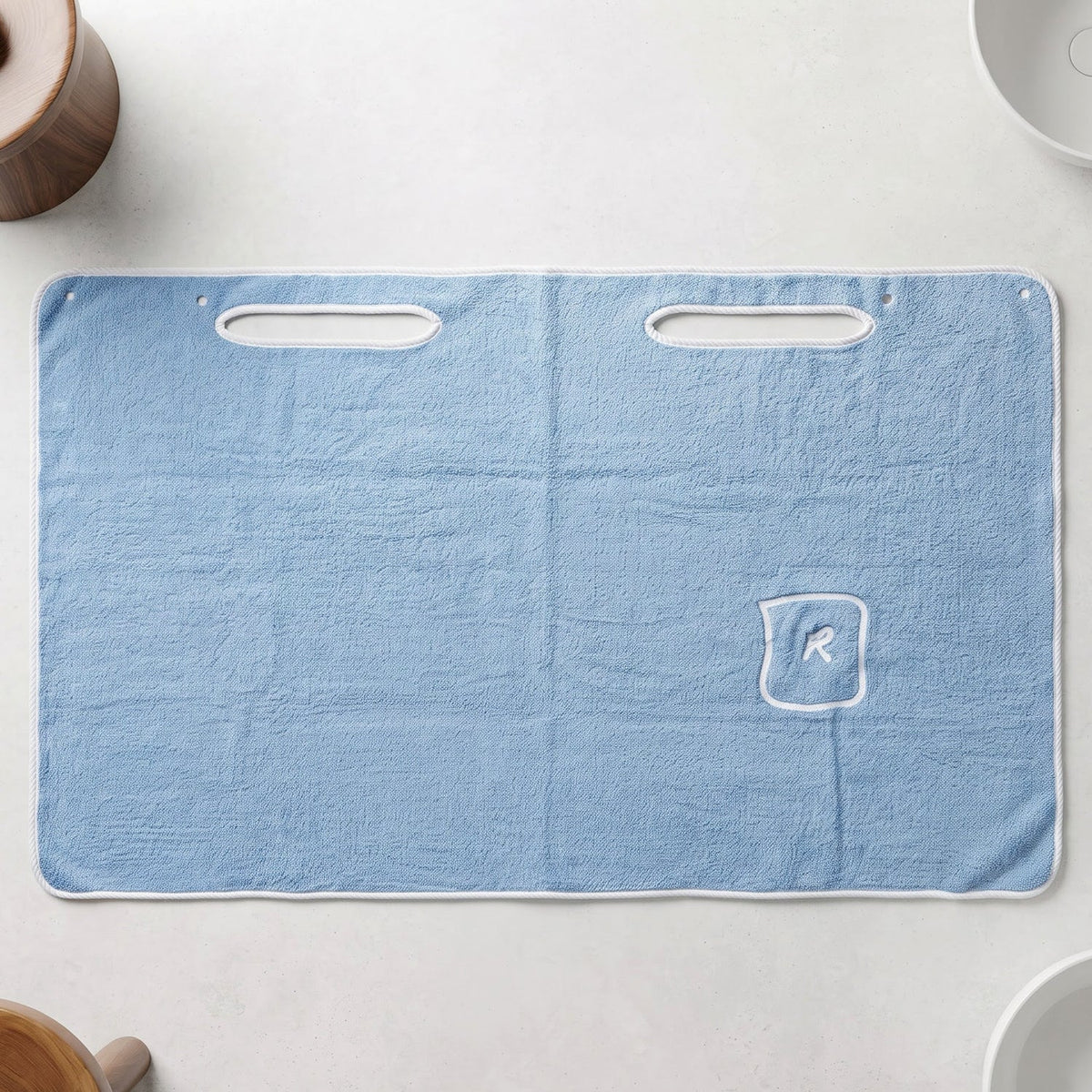 Flat lay of a light blue rectangular bath towel wrap with two armholes and a small pocket with an R monogram.