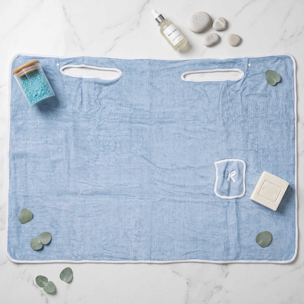 Flat lay of a light blue bath towel wrap surrounded by bath salts, essential oil, soap, and eucalyptus leaves.