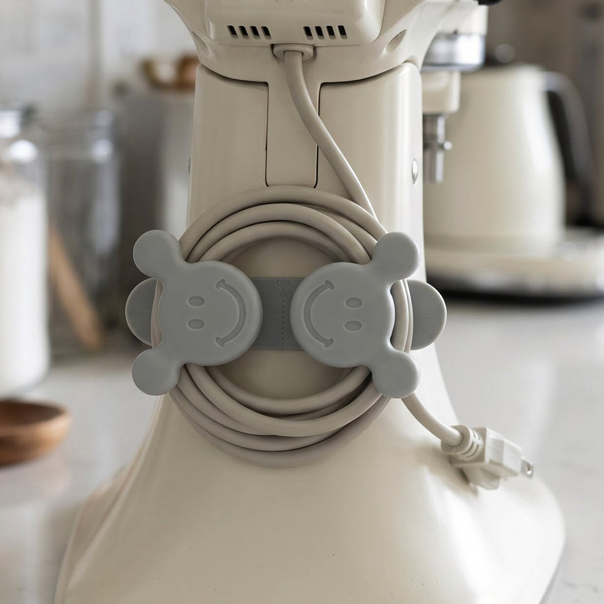 A gray silicone cord organizer with two smiling faces, holding a coiled light gray appliance cord on a white stand mixer.