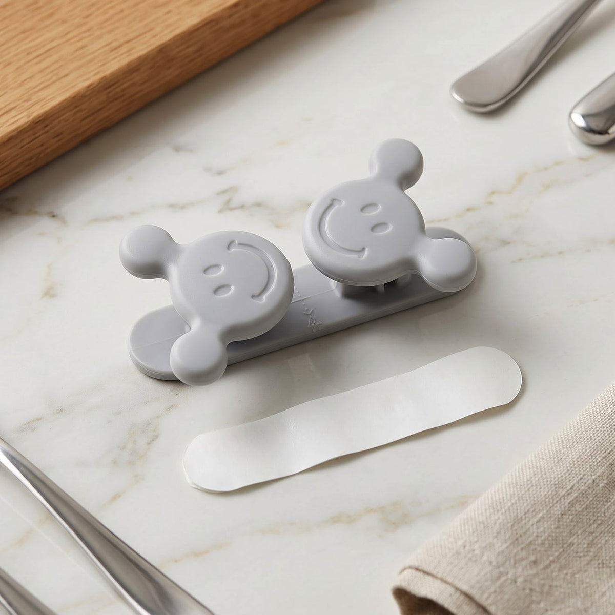 A gray silicone cord organizer with two smiling faces and a white adhesive strip on a marble countertop next to silverware.