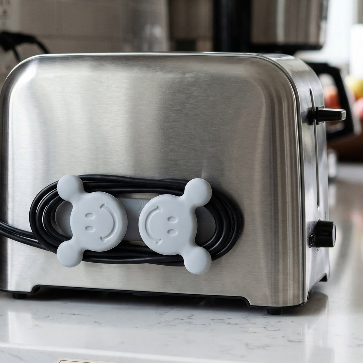 A gray silicone cord organizer with two smiling faces, holding a coiled black appliance cord on a stainless steel toaster.