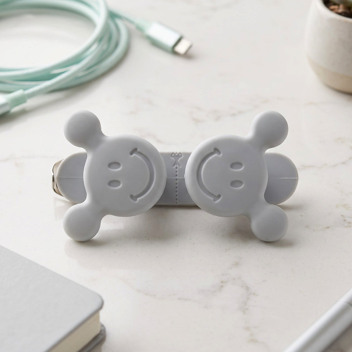 A gray silicone cord organizer with two smiling faces on a marble surface, surrounded by a light blue cable and a small