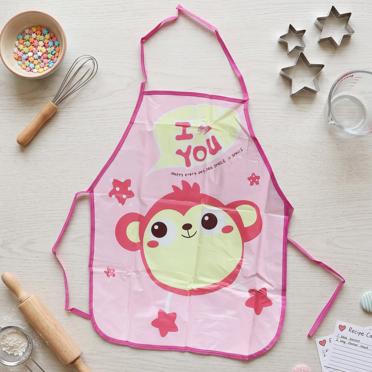 A pink monkey print apron is laid flat on a white wooden surface, surrounded by baking ingredients and tools.