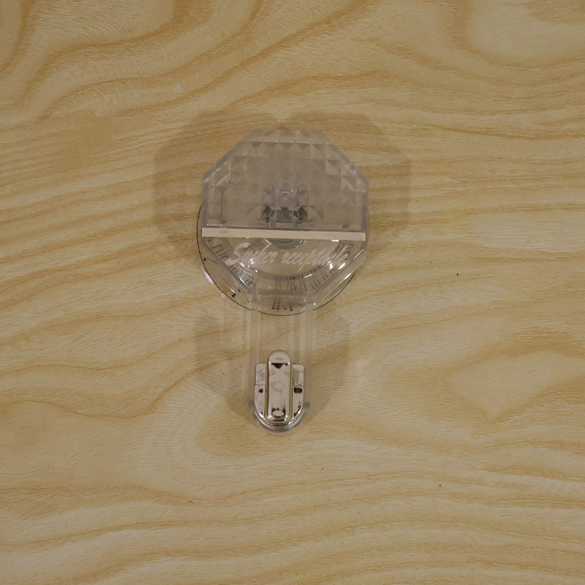 A clear, diamond-patterned self-adhesive hook with a silver hook, attached to a light wooden surface.