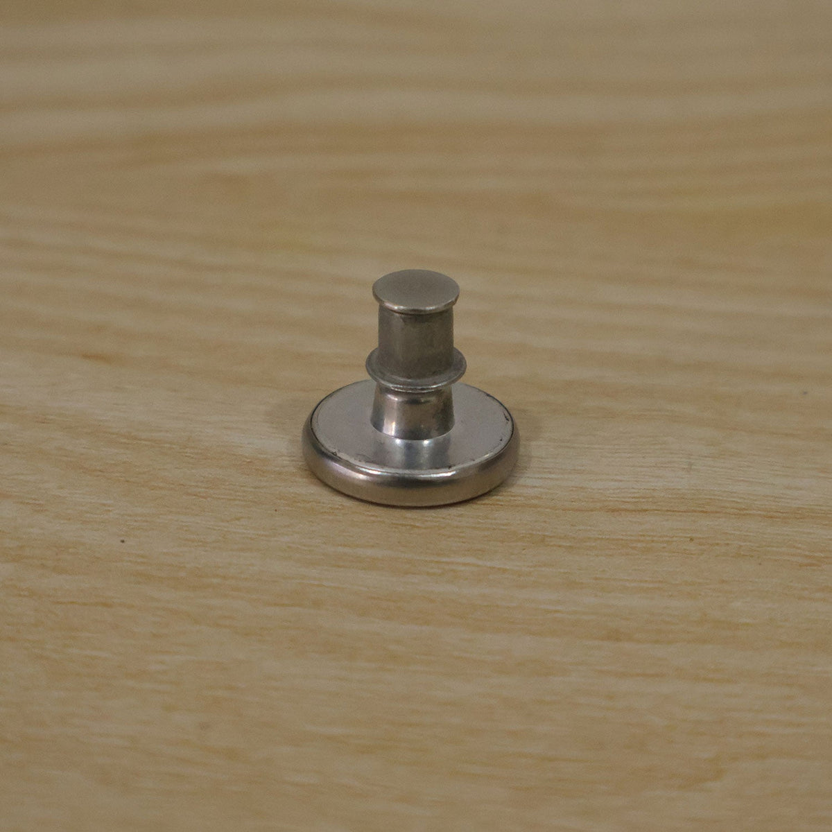 A silver metal button pin with its back lock attached, sitting on a light wooden surface.