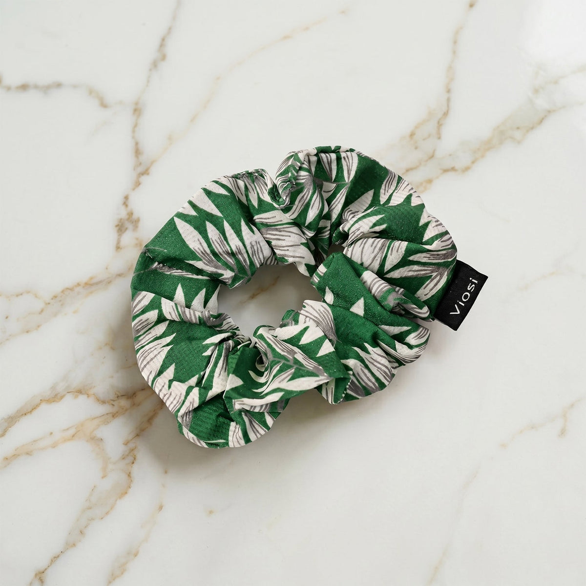 A green and white leaf print scrunchie with a black 'viosi' tag is displayed on a white and grey marble background.