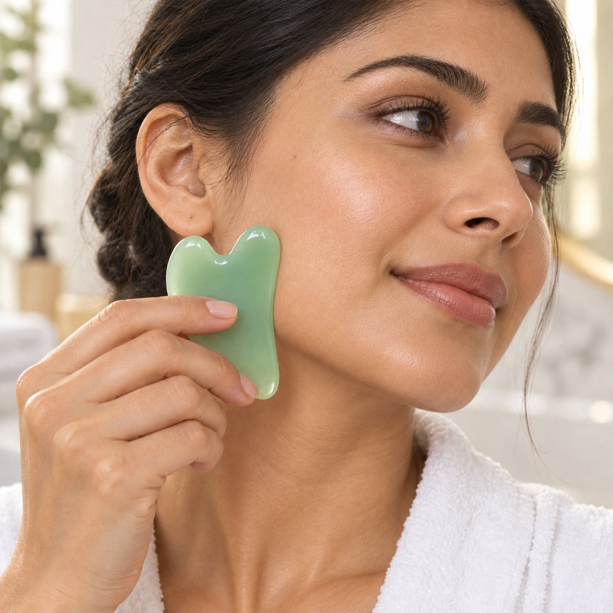 A woman gently uses a green gua sha tool on her cheek, looking away with a slight smile.