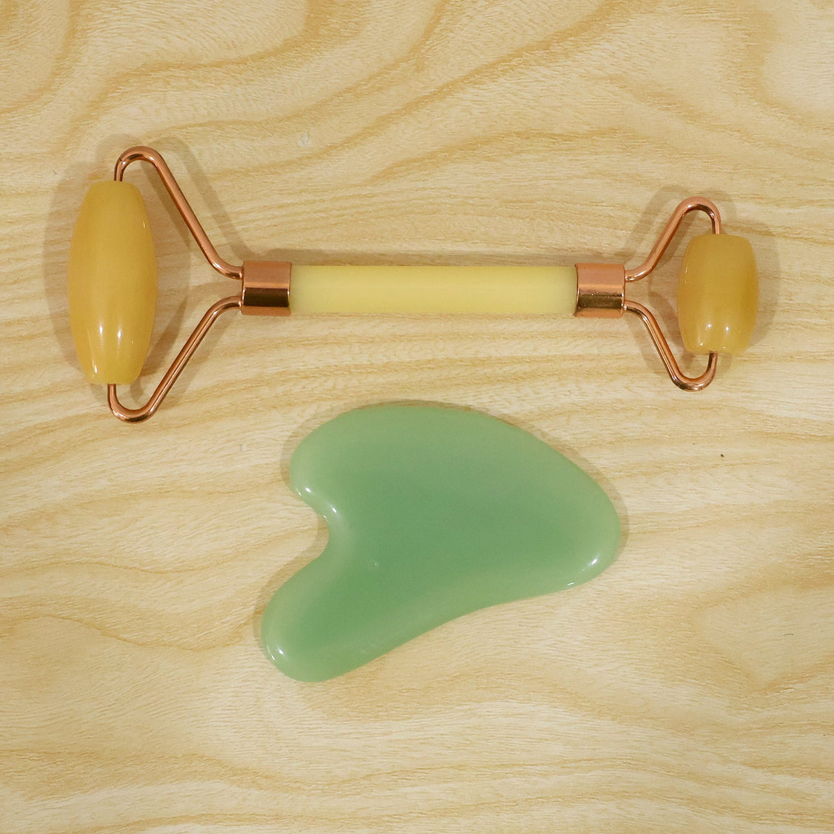 A yellow facial roller with rose gold accents and a green gua sha tool are positioned on a light wooden surface.