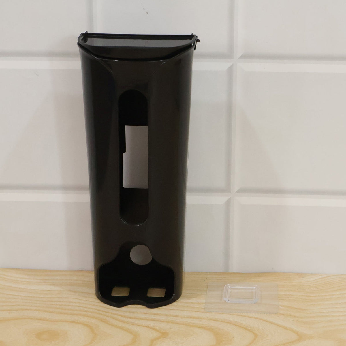 A black plastic bag dispenser sits on a wooden surface next to a clear adhesive hook on a white tiled wall.