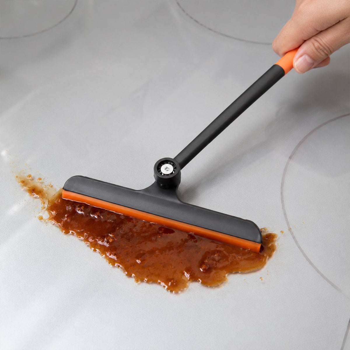 A hand uses an orange and black squeegee to clean a brown liquid spill on a light gray surface.