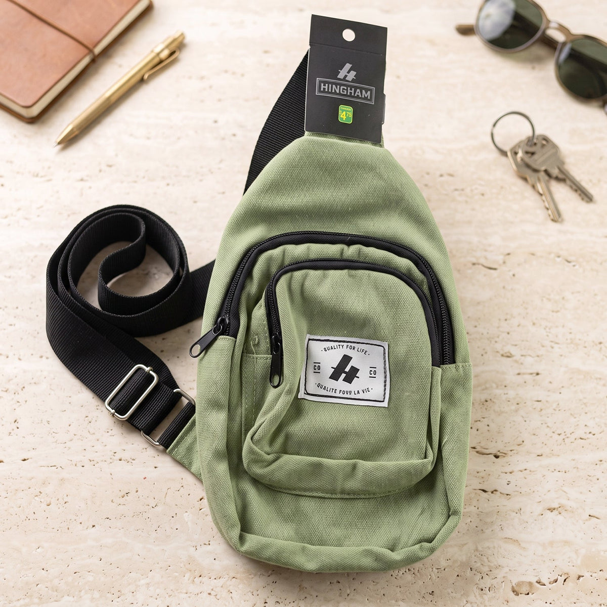 A green canvas sling bag with a black strap and zippers, lying on a light-colored textured surface with a pen, keys, and