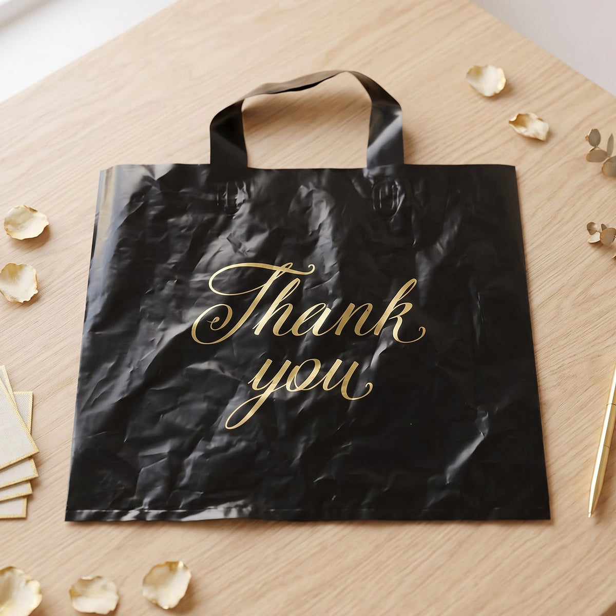 A black plastic 'Thank You' bag with gold script is laid flat on a wooden surface with scattered rose petals.