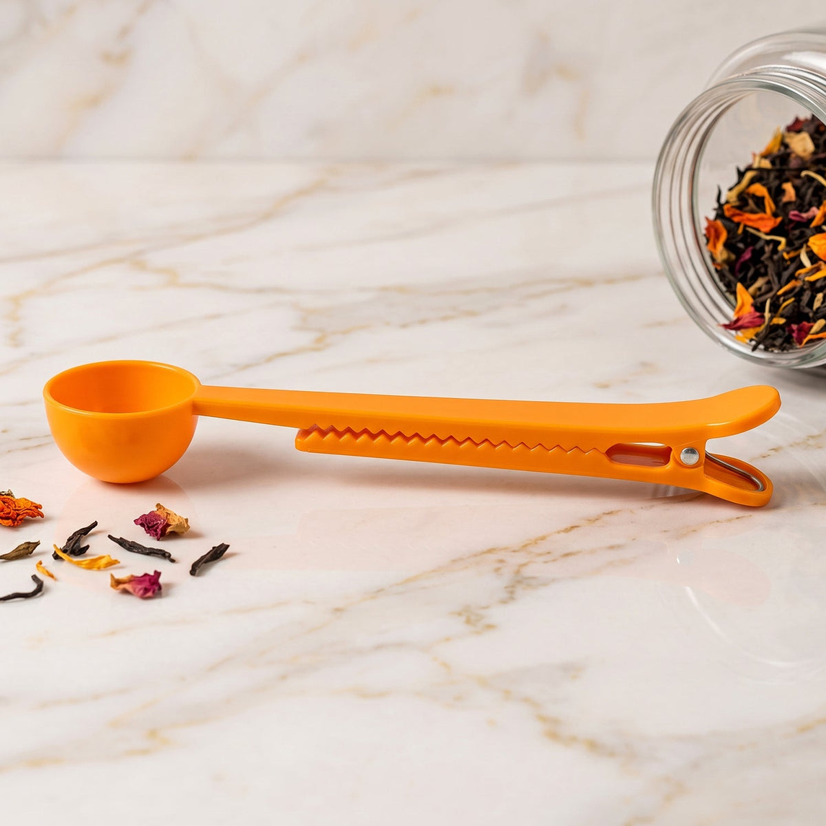 An orange plastic scoop and clip lies on a marble surface, with loose tea leaves scattered around and a jar of tea.