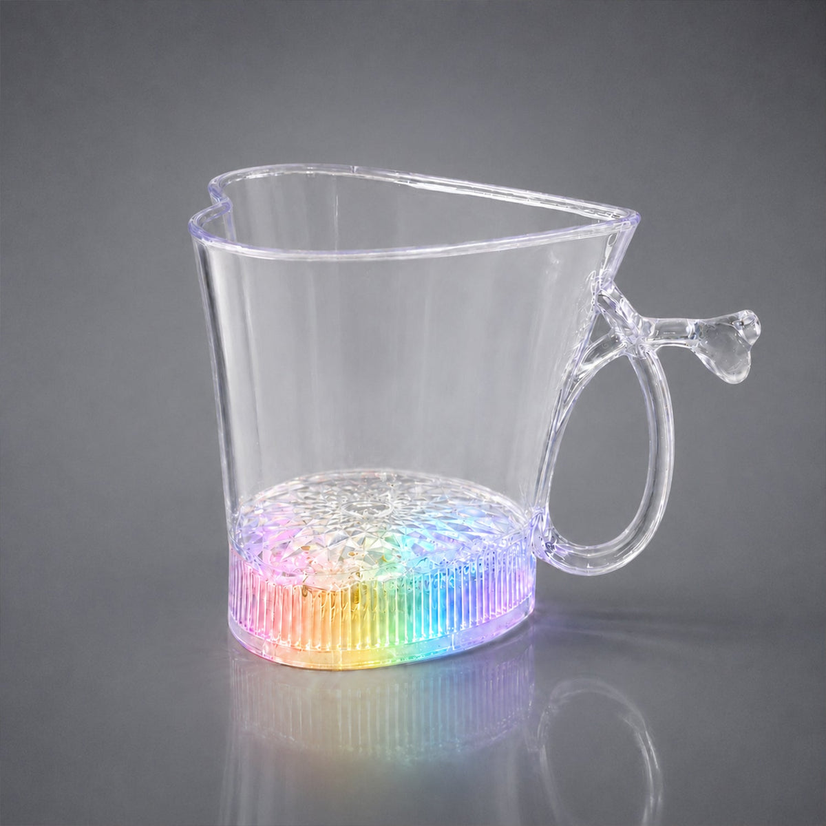 A clear heart-shaped mug with a rainbow-lit base, reflecting on a gray surface.